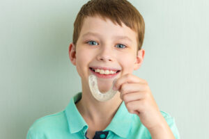 Smiling teenage boy holding aligners, silicone cap for teeth straightening. Orthodontist correcting bite.