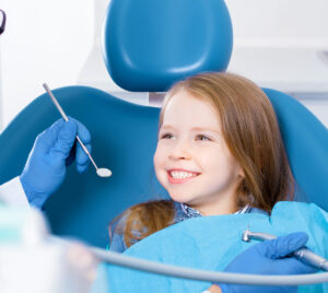 Little cute smiling girl is sitting in dental chair and opening mouth in clinic, office. Doctor is preparing for examination of child teeth with tools, instruments. Visiting dentist with children.