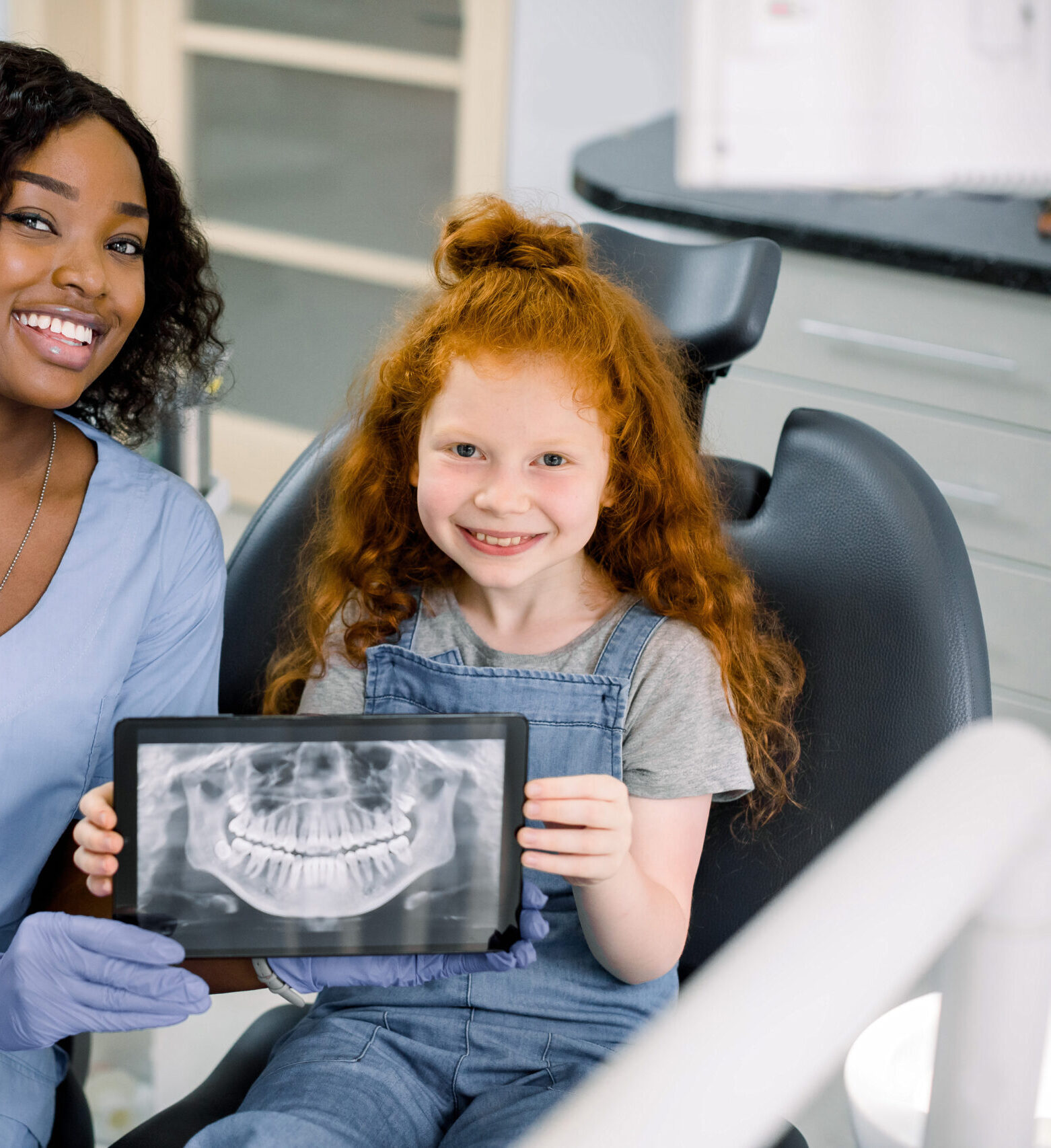 Little smiling girl with red curly hair sitting on chair and looking at camera, while holding x-ray scan image of her teeth on digital tablet together with her cheerful black female dentist at clinic.