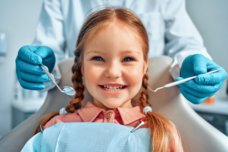 Cropped portrait of little girl with pigtails hair sitting in dental chair looking at camera and smiling. Behind, a doctor in gloves holds examination tools.Children's dentistry.